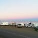 Camper e camper sono parcheggiati lungo una strada costiera al crepuscolo. Le persone stanno vicino alla costa e sono presenti diversi veicoli, tra cui un'auto della polizia. Il cielo passa dal rosa al blu man mano che la luce del giorno svanisce. Il mare calmo si estende fino all'orizzonte.