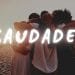 Quattro persone che si abbracciano su una spiaggia durante il tramonto. La parola "SAUDADE" è ben visibile in grassetto bianco sull'immagine.