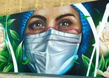 Un murale raffigura il primo piano di una donna che indossa un berretto chirurgico blu e una maschera bianca. I suoi occhi espressivi suggeriscono un'espressione seria. Intorno a lei ci sono rose bianche e foglie verdi che aggiungono un tocco di natura all'opera d'arte.