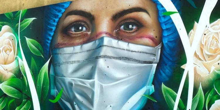 Un murale raffigura il primo piano di una donna che indossa un berretto chirurgico blu e una maschera bianca. I suoi occhi espressivi suggeriscono un'espressione seria. Intorno a lei ci sono rose bianche e foglie verdi che aggiungono un tocco di natura all'opera d'arte.