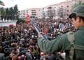 Immagine scattata in Portogallo 25 aprile del 1978. Un soldato con in mano un fucile ornato da un garofano rosso si trova su un tetto che domina una grande folla riunita in una pubblica piazza. La scena sembra essere una protesta o manifestazione pacifica. Sullo sfondo sono visibili edifici residenziali e alberi.