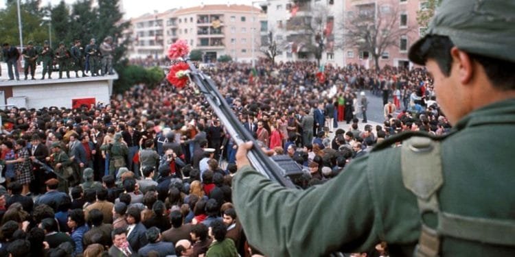 Immagine scattata in Portogallo 25 aprile del 1978. Un soldato con in mano un fucile ornato da un garofano rosso si trova su un tetto che domina una grande folla riunita in una pubblica piazza. La scena sembra essere una protesta o manifestazione pacifica. Sullo sfondo sono visibili edifici residenziali e alberi.