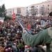 Immagine scattata in Portogallo 25 aprile del 1978. Un soldato con in mano un fucile ornato da un garofano rosso si trova su un tetto che domina una grande folla riunita in una pubblica piazza. La scena sembra essere una protesta o manifestazione pacifica. Sullo sfondo sono visibili edifici residenziali e alberi.