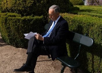 Un uomo in abito scuro e cravatta blu siede su una panchina verde in un giardino ben curato, leggendo e scrivendo sui fogli che ha in mano. La luce del sole illumina la scena, proiettando ombre chiare sul sentiero di ghiaia che circonda le rigogliose siepi verdi.