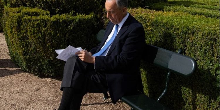 Un uomo in abito scuro e cravatta blu siede su una panchina verde in un giardino ben curato, leggendo e scrivendo sui fogli che ha in mano. La luce del sole illumina la scena, proiettando ombre chiare sul sentiero di ghiaia che circonda le rigogliose siepi verdi.