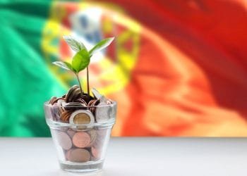 Una piccola pianta con foglie verdi cresce da un bicchiere pieno di monete, a simboleggiare la crescita finanziaria e gli investimenti. Lo sfondo presenta un'immagine sfocata della bandiera portoghese, con i colori rosso, verde e giallo visibili.