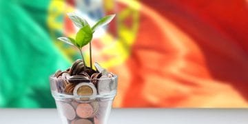 Una piccola pianta con foglie verdi cresce da un bicchiere pieno di monete, a simboleggiare la crescita finanziaria e gli investimenti. Lo sfondo presenta un'immagine sfocata della bandiera portoghese, con i colori rosso, verde e giallo visibili.