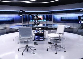 Un moderno studio televisivo con un tavolo rotondo in vetro e due sedie da ufficio una di fronte all'altra. Lo sfondo è caratterizzato da grandi schermi digitali e apparecchiature di illuminazione professionali. Sopra è visibile una gru per telecamere e i monitor sono posizionati sotto il tavolo.