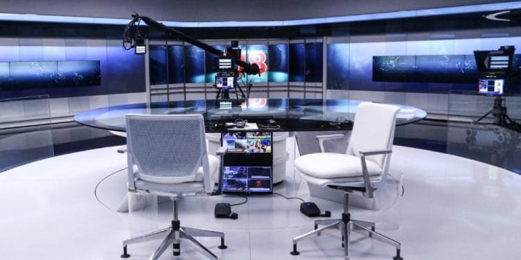 Un moderno studio televisivo con un tavolo rotondo in vetro e due sedie da ufficio una di fronte all'altra. Lo sfondo è caratterizzato da grandi schermi digitali e apparecchiature di illuminazione professionali. Sopra è visibile una gru per telecamere e i monitor sono posizionati sotto il tavolo.