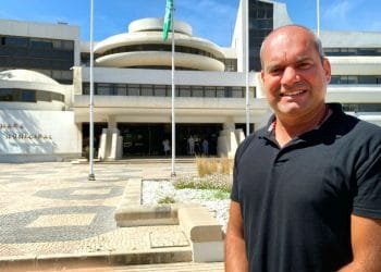 Un uomo sorridente che indossa una camicia nera si trova di fronte a un edificio moderno e curvo su cui è scritta la scritta "Câmara Municipal". L'edificio presenta pareti esterne bianche, un cortile piastrellato a motivi geometrici, diverse aste per bandiere e un cielo azzurro e limpido sopra.