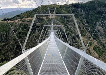 Un lungo ponte sospeso in metallo si estende su un profondo canyon boscoso con uno sfondo di aspre montagne e cieli nuvolosi. Il ponte, realizzato in grata di acciaio, ha telai alti sostenuti da cavi che si aggiungono al suo design architettonico moderno.