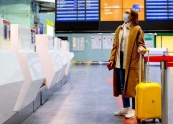 Una donna che indossa una maschera, un lungo cappotto marrone e occhiali si trova in un aeroporto vicino al banco del check-in con una valigia giallo brillante. Alza lo sguardo verso un pannello informativo che mostra i dettagli del volo.