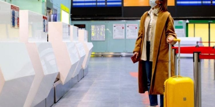 Una donna che indossa una maschera, un lungo cappotto marrone e occhiali si trova in un aeroporto vicino al banco del check-in con una valigia giallo brillante. Alza lo sguardo verso un pannello informativo che mostra i dettagli del volo.