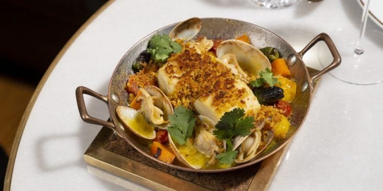 Una padella di metallo su un tavolo bianco contiene un piatto gourmet di frutti di mare a base di vongole, pesce e verdure guarniti con coriandolo fresco. Sullo sfondo sono visibili un bicchiere di vino bianco e un bicchiere d'acqua con intricati dettagli.
