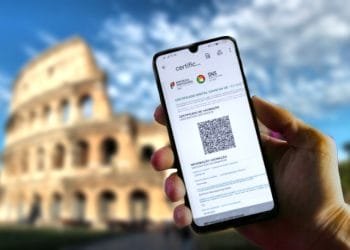 Una mano che tiene uno smartphone su cui è visualizzato un certificato di vaccinazione digitale con un codice QR davanti all'iconico Colosseo di Roma in una giornata soleggiata con cielo parzialmente nuvoloso.