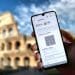 Una mano che tiene uno smartphone su cui è visualizzato un certificato di vaccinazione digitale con un codice QR davanti all'iconico Colosseo di Roma in una giornata soleggiata con cielo parzialmente nuvoloso.