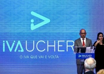 Un uomo in abito grigio e cravatta rossa è in piedi su un podio con il logo "IVAUCHER" visualizzato a grandi lettere su uno schermo blu dietro di lui. Alla sua sinistra, una donna sta interpretando il linguaggio dei segni. Sotto il logo è visibile la tagline "O IVA QUE VAI E VOLTA".