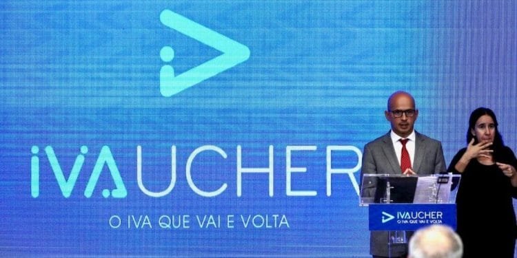 Un uomo in abito grigio e cravatta rossa è in piedi su un podio con il logo "IVAUCHER" visualizzato a grandi lettere su uno schermo blu dietro di lui. Alla sua sinistra, una donna sta interpretando il linguaggio dei segni. Sotto il logo è visibile la tagline "O IVA QUE VAI E VOLTA".
