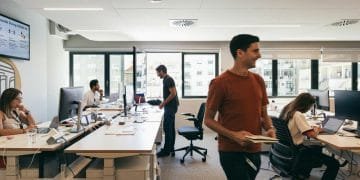 Un ufficio moderno con diverse persone che lavorano su scrivanie dotate di computer. Una persona con una maglietta rossa e un taccuino in mano passa, mentre gli altri sono seduti e concentrati sui loro schermi. Le grandi finestre sullo sfondo lasciano entrare la luce naturale.