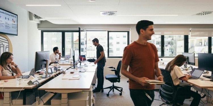 Un ufficio moderno con diverse persone che lavorano su scrivanie dotate di computer. Una persona con una maglietta rossa e un taccuino in mano passa, mentre gli altri sono seduti e concentrati sui loro schermi. Le grandi finestre sullo sfondo lasciano entrare la luce naturale.