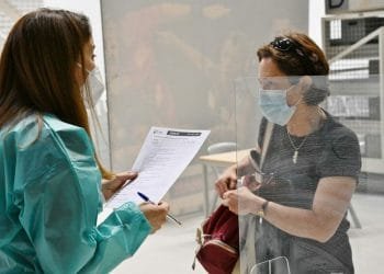 Un operatore sanitario in abito medico verde acqua e maschera tiene in mano un blocco per appunti e parla con una donna in camicia nera e maschera a un tavolo diviso. La donna stringe una borsa rossa e sta dietro una barriera trasparente.
