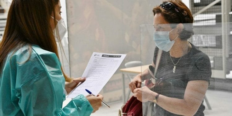 Un operatore sanitario in abito medico verde acqua e maschera tiene in mano un blocco per appunti e parla con una donna in camicia nera e maschera a un tavolo diviso. La donna stringe una borsa rossa e sta dietro una barriera trasparente.