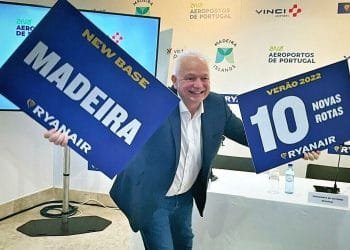 Un uomo in giacca e cravatta sorride e tiene in mano due cartelli. Il cartello nella sua mano destra recita "NEW BASE MADEIRA RYANAIR" e il cartello nella sua mano sinistra recita "VERÃO 2022 10 NOVAS ROTAS RYANAIR". Si trova davanti a uno sfondo con loghi e testi sugli aeroporti di Vinci e sugli aeroporti del Portogallo, annunciando che Ryanair vola a Madeira
