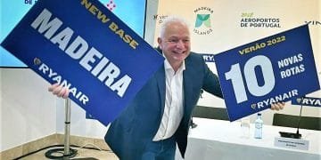 Un uomo in giacca e cravatta sorride e tiene in mano due cartelli. Il cartello nella sua mano destra recita "NEW BASE MADEIRA RYANAIR" e il cartello nella sua mano sinistra recita "VERÃO 2022 10 NOVAS ROTAS RYANAIR". Si trova davanti a uno sfondo con loghi e testi sugli aeroporti di Vinci e sugli aeroporti del Portogallo, annunciando che Ryanair vola a Madeira