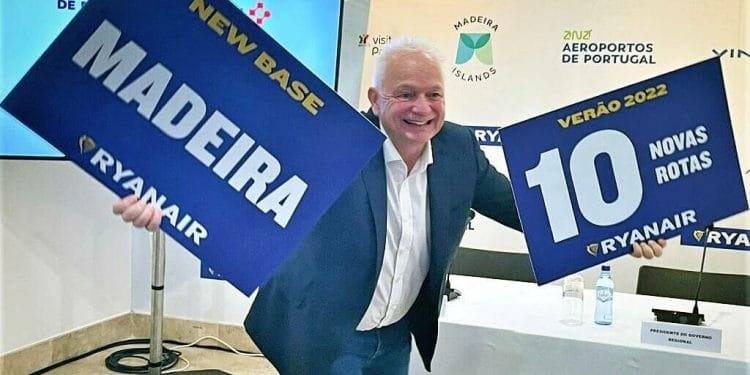 Un uomo in giacca e cravatta sorride e tiene in mano due cartelli. Il cartello nella sua mano destra recita "NEW BASE MADEIRA RYANAIR" e il cartello nella sua mano sinistra recita "VERÃO 2022 10 NOVAS ROTAS RYANAIR". Si trova davanti a uno sfondo con loghi e testi sugli aeroporti di Vinci e sugli aeroporti del Portogallo, annunciando che Ryanair vola a Madeira