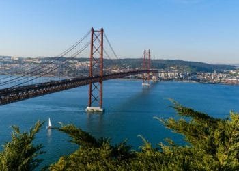 Un grande ponte sospeso rosso attraversa un ampio fiume blu e collega due aree urbane. Una piccola barca a vela galleggia sotto il ponte. Il fogliame verde è visibile in primo piano e sia il cielo che l'acqua sono limpidi e blu.