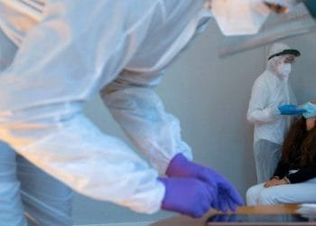 Gli operatori sanitari in tute protettive per tutto il corpo, visiere e maschere stanno somministrando o preparando i test COVID-19. Un operatore sta tamponando il naso di un paziente seduto, mentre un altro è concentrato su un tablet o un'attrezzatura.