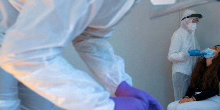 Gli operatori sanitari in tute protettive per tutto il corpo, visiere e maschere stanno somministrando o preparando i test COVID-19. Un operatore sta tamponando il naso di un paziente seduto, mentre un altro è concentrato su un tablet o un'attrezzatura.