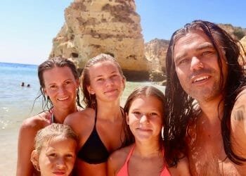 Una famiglia Bitcoin di cinque persone, tra cui due adulti e tre bambini, si fa un selfie su una spiaggia soleggiata in Portogallo con acque cristalline e scogliere rocciose sullo sfondo. Tutti sorridono e indossano costumi da bagno, godendosi una giornata perfetta in spiaggia.