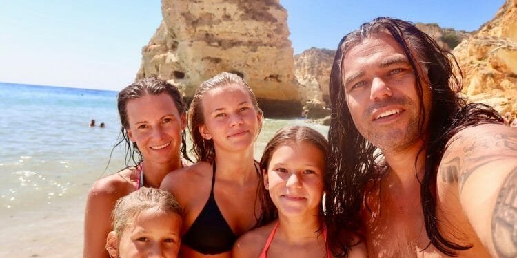 Una famiglia Bitcoin di cinque persone, tra cui due adulti e tre bambini, si fa un selfie su una spiaggia soleggiata in Portogallo con acque cristalline e scogliere rocciose sullo sfondo. Tutti sorridono e indossano costumi da bagno, godendosi una giornata perfetta in spiaggia.