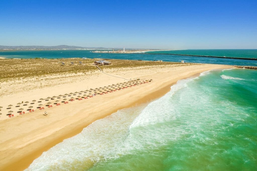 Veduta aerea di una spiaggia sabbiosa con file di ombrelloni e sedie a sdraio in Algarve. L'acqua limpida e turchese lambisce dolcemente la riva, mentre sullo sfondo si possono vedere la vegetazione rada e le montagne. Un molo si estende nell'acqua sul lato destro. Giornata luminosa e soleggiata con cielo terso, che cattura la serena bellezza dell'autunno.