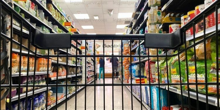 Una prospettiva dall'interno di un carrello della spesa mentre viene spinto lungo la corsia del supermercato. Gli scaffali su entrambi i lati sono riforniti di vari prodotti, compreso il cibo per animali domestici. Il negozio è ben illuminato e in lontananza, alla fine del corridoio, si vede una persona che sta valutando i costi del viaggio in Portogallo.