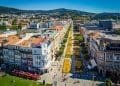 Una vivace veduta aerea di una città con una lunga strada pedonale fiancheggiata da fiori fiancheggiata da edifici storici con tetti di tegole rosse. Uno scenario scenografico di colline si estende in lontananza. Fiori gialli brillanti dominano il percorso centrale e si vedono persone passeggiare: una scena perfetta per investire in Portogallo.