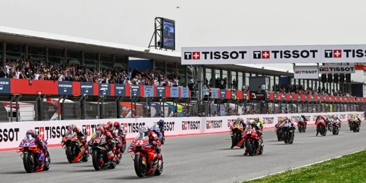 I motociclisti sfrecciano lungo la pista dell'Autódromo Internacional do Algarve, circondati dagli spettatori sugli spalti. Il circuito presenta il marchio Tissot su striscioni e strutture. I motociclisti indossano equipaggiamento colorato e la pista è fiancheggiata da erba.