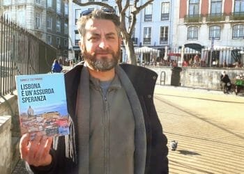 Un uomo con i capelli corti e la barba tiene in mano un libro intitolato "Lisbona è un'assurda speranza" di Daniele Coltrinari mentre si trova all'aperto in una soleggiata area urbana. Sullo sfondo si vedono edifici e un albero, insieme a una piccola folla di persone.