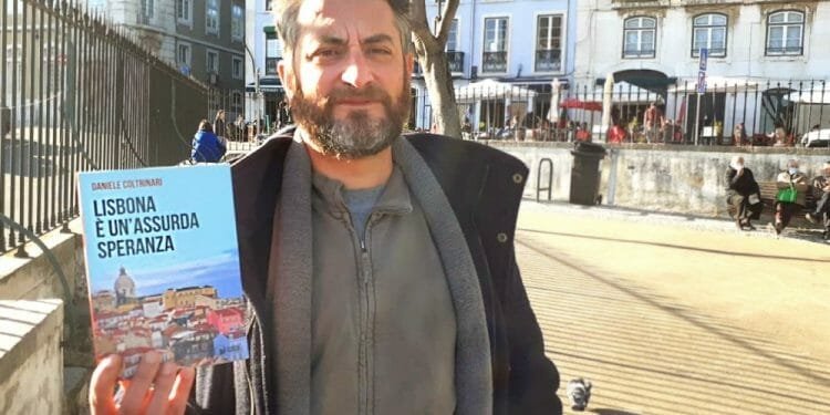 Un uomo con i capelli corti e la barba tiene in mano un libro intitolato "Lisbona è un'assurda speranza" di Daniele Coltrinari mentre si trova all'aperto in una soleggiata area urbana. Sullo sfondo si vedono edifici e un albero, insieme a una piccola folla di persone.
