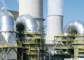 Complesso industriale con grandi condutture metalliche e torri di raffreddamento. Le strutture sono collegate da passerelle con ringhiera gialla, mentre il vapore fuoriesce dalle torri di raffreddamento cilindriche, indicando il funzionamento attivo. Questo sito, che ricorda gli impianti di Enel Portogallo, incarna l'efficienza industriale.