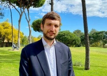 Un uomo con i capelli castano chiaro e la barba, che indossa una giacca scura sopra una camicia bianca, sta in piedi sul prato con alberi ad alto fusto. C'è qualcuno seduto su una panchina sullo sfondo e il cielo è sereno con qualche nuvola. Massimo Ungaro del COMITES potrebbe facilmente far parte di questa scena serena.