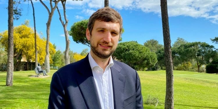 Un uomo con i capelli castano chiaro e la barba, che indossa una giacca scura sopra una camicia bianca, sta in piedi sul prato con alberi ad alto fusto. C'è qualcuno seduto su una panchina sullo sfondo e il cielo è sereno con qualche nuvola. Massimo Ungaro del COMITES potrebbe facilmente far parte di questa scena serena.