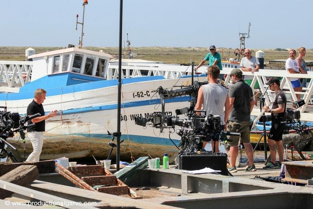 Una troupe cinematografica sta girando una scena in un cantiere navale con un peschereccio bianco e blu sullo sfondo. Il regista gesticola con le mani mentre la troupe aziona le telecamere e l'attrezzatura audio. Gli spettatori guardano da dietro la barca, curiosi se è per i prossimi World Trailer Awards.