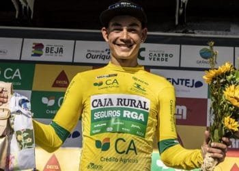 Un ciclista in maglia gialloverde della "Caja Rural" sorride e tiene in una mano un mazzo di girasoli e nell'altra una bottiglia di champagne, festeggiando la sua vittoria alla Volta ao Alentejo. Lo sfondo presenta vari loghi degli sponsor.