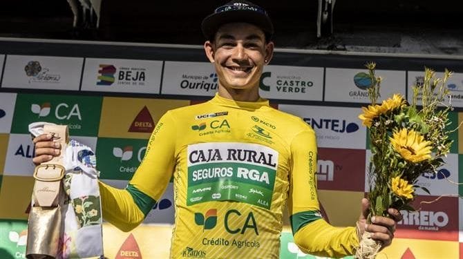 Un ciclista in maglia gialloverde della "Caja Rural" sorride e tiene in una mano un mazzo di girasoli e nell'altra una bottiglia di champagne, festeggiando la sua vittoria alla Volta ao Alentejo. Lo sfondo presenta vari loghi degli sponsor.