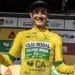 Un ciclista in maglia gialloverde della "Caja Rural" sorride e tiene in una mano un mazzo di girasoli e nell'altra una bottiglia di champagne, festeggiando la sua vittoria alla Volta ao Alentejo. Lo sfondo presenta vari loghi degli sponsor.