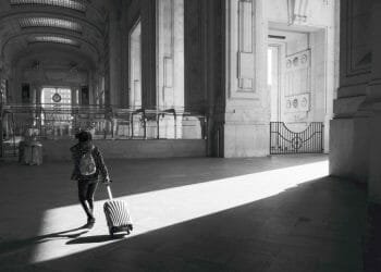 Una persona che indossa uno zaino trascina una valigia attraverso una grande stazione ferroviaria illuminata dal sole con grandi caratteristiche architettoniche e alti soffitti ad arco. La luce proietta lunghe ombre sul pavimento, evidenziando l'ampio spazio: una toccante scena di emigrazione.