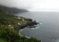 Un paesaggio costiero caratterizzato da piccole case bianche dai tetti rossi sparse sulle verdi scogliere dell'Ilha de São Jorge, a picco sul mare. Le onde dell'oceano si infrangono contro gli affioramenti rocciosi scuri, mentre la nebbia e le nuvole coprono parzialmente le montagne sullo sfondo.