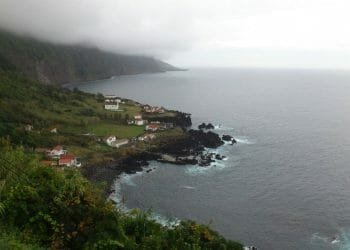 Un paesaggio costiero caratterizzato da piccole case bianche dai tetti rossi sparse sulle verdi scogliere dell'Ilha de São Jorge, a picco sul mare. Le onde dell'oceano si infrangono contro gli affioramenti rocciosi scuri, mentre la nebbia e le nuvole coprono parzialmente le montagne sullo sfondo.